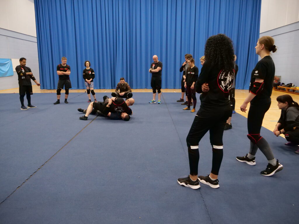 A group of people practicing krav maga are standing in a gymnasium.
