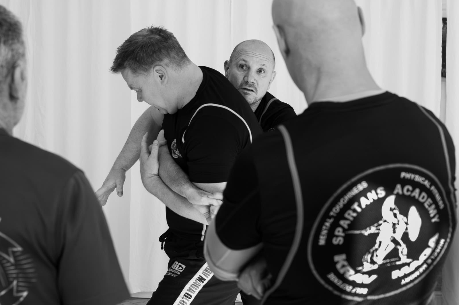 Four men are in a self-defense training session, with one man demonstrating a grappling or restraint technique on another, while two others watch.