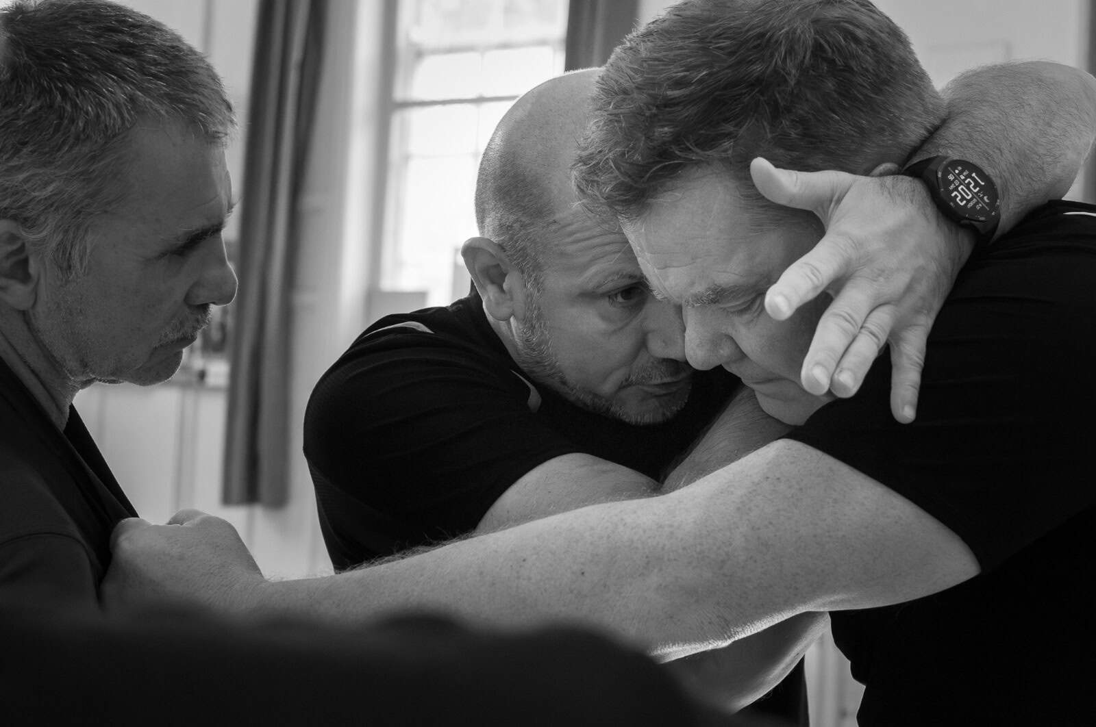 Three men practicing a close-contact self-defense or martial arts technique indoors, focusing intently on their movements.