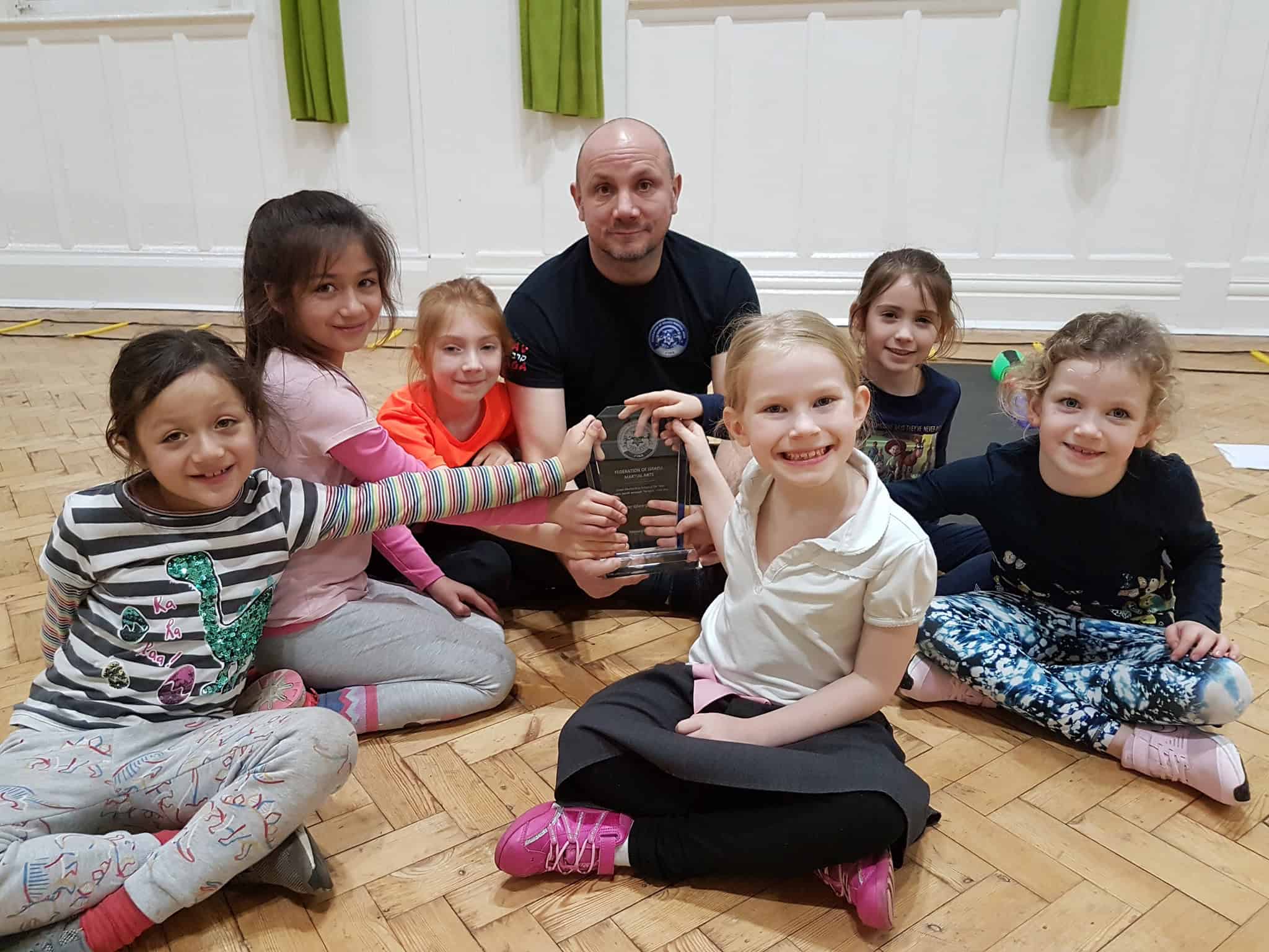 A group of seven people, six children and one adult, sit on a wooden floor indoors. The smiling children hold an award or trophy together, celebrating their achievements in krav maga self defence training.